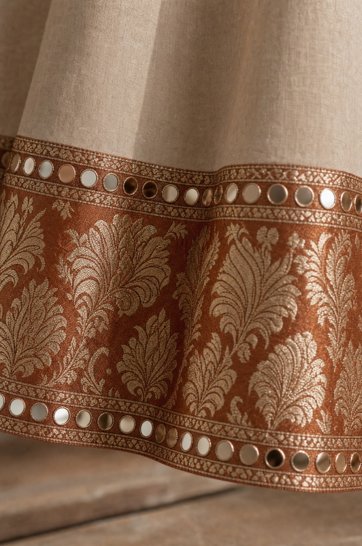 Close view of a hem border in copper brocade, edged above and below with rows of round mirrors in gold rings.